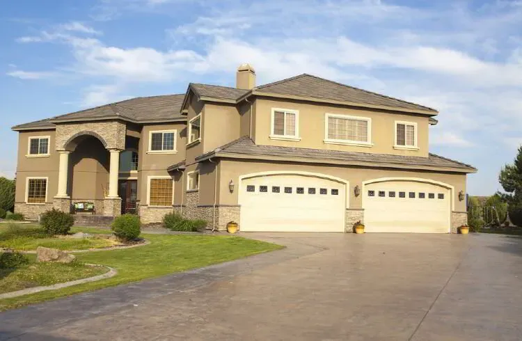Tan stucco two-story home with cream garage doors and a stone archway after an exterior repaint by Elegant Edge Painting.